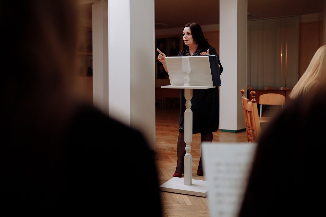 A woman conductor instructing a music class indoors, pointing and presenting.